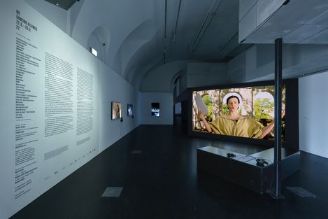 Dark exhibition space with white wall displaying text and multiple screens showing video installations