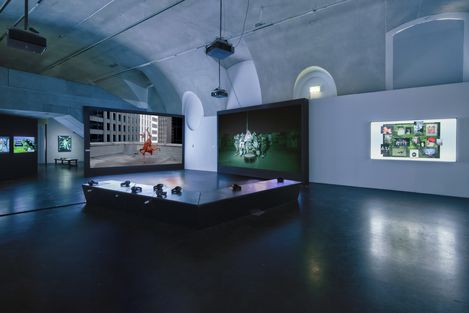 Dimly lit exhibition space with multiple large screens showing videos and photos and a platform with headphones in the center