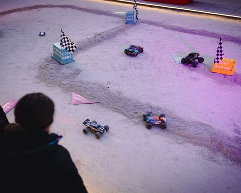 Vier ferngesteuerte Autos fahren auf einer sandigen Rennstrecke mit kleinen Flaggen und Hindernissen, zwei Personen beobachten das Geschehen im Vordergrund