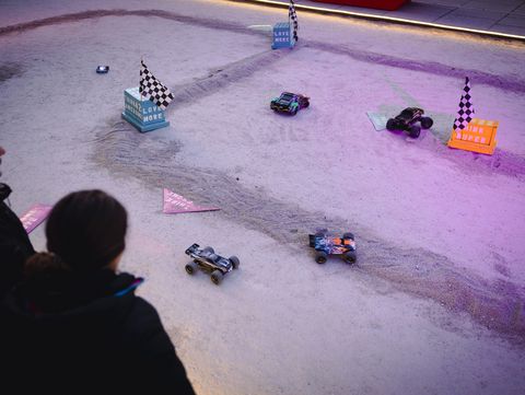 Vier ferngesteuerte Autos fahren auf einer sandigen Rennstrecke mit kleinen Flaggen und Hindernissen, zwei Personen beobachten das Geschehen im Vordergrund