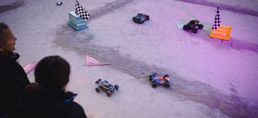 Four remote-controlled cars driving on a sandy race track with small flags and obstacles, two people watching in the foreground