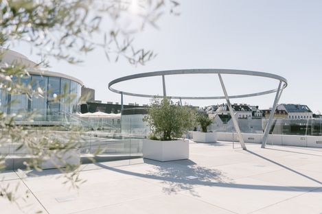 Sunny rooftop terrace of a modern building with a curved glass façade, spacious planters, and a striking sculptural circular light feature supported by angled columns. Olive branches frame the architecture in the foreground.