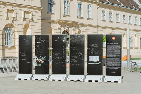 Six black information panels featuring white handwriting, drawings, and photos are located in front of the MQ building