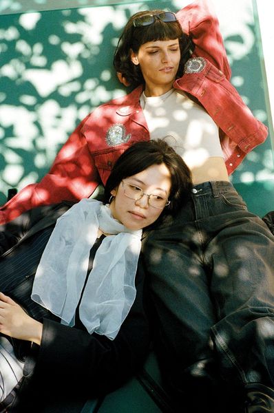 Two women lying relaxed next to each other on a green surface, one wearing a red jacket with sunglasses on her head, the other wearing black clothing with a white sheer scarf and glasses.