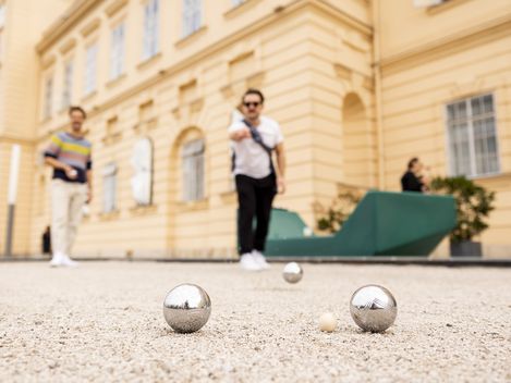Zwei Personen werfen Boulekugeln in den Sand der Boulebahn am Vorplatz des MQ. Im Hintergrunde in grünblaues Enzi.