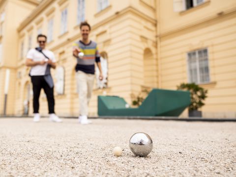 Two men playing boule in a sandy courtyard. One throws a metal ball towards a wooden target ball. They appear relaxed and focused.
