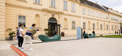 Two men playing boule in a sandy courtyard. One throws a metal ball towards a wooden target ball. They appear relaxed and focused.