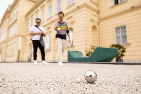 Two men playing boule in a sandy courtyard. One throws a metal ball towards a wooden target ball. They appear relaxed and focused.