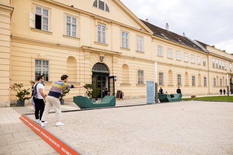 Two men playing boule in a sandy courtyard. One throws a metal ball towards a wooden target ball. They appear relaxed and focused.
