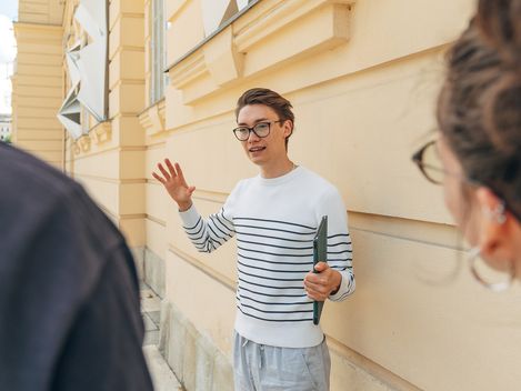 Eine Person in einem gestreiften Pullover steht vor der gelben Fassade des MuseumsQuartier Wien mit weißen, dreieckigen Wanddekorationen und spricht zu zwei anderen Personen