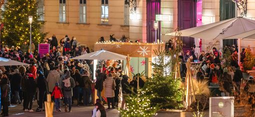 Crowd of people at a Christmas market in front of a historic building with lit windows and Christmas trees.