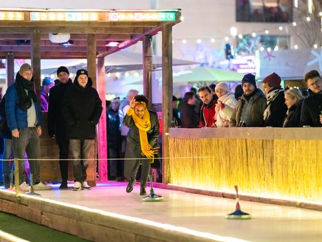 Eisstockbahn, Winter im MQ. Menschen spielen Eisstock. Eine Person wirft  begeistert einen Curlingstein auf die Eisstockbahn im MQ Hof, beobachtet von einer Menschenmenge, die in Wintermäntel gehüllt ist.