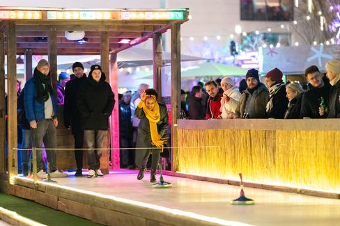 Eisstockbahn, Winter im MQ. Menschen spielen Eisstock. Eine Person wirft  begeistert einen Curlingstein auf die Eisstockbahn im MQ Hof, beobachtet von einer Menschenmenge, die in Wintermäntel gehüllt ist.