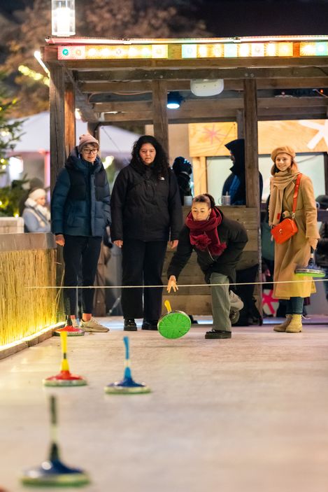 Vier Personen stehen auf einer beleuchteten Bahn, eine Person wirft einen Curlingstein, die anderen beobachten.