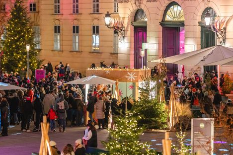 Menschenmenge auf einem Weihnachtsmarkt vor einem historischen Gebäude mit beleuchteten Weihnachtsbäumen und Ständen