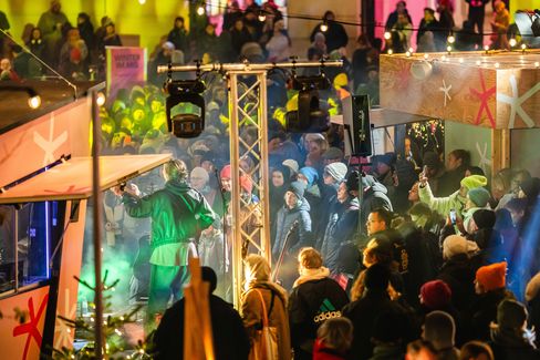 Menschenmenge bei einer beleuchteten Veranstaltung im Freien mit einer Person auf einer Bühne und Lichterketten im Hintergrund