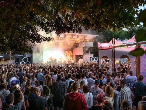 Große Menschenmenge vor einer Bühne bei einem Open-Air-Konzert unter Bäumen am Abend.