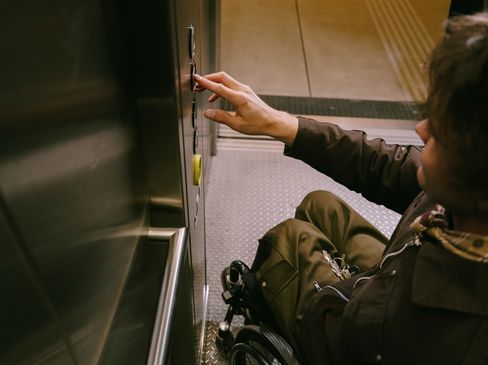 Eine Person im Rollstuhl betätigt die Bedienelemente eines barrierefreien Aufzugs. Die Aufnahme zeigt den Innenraum des Aufzugs mit gut erreichbaren Tasten.