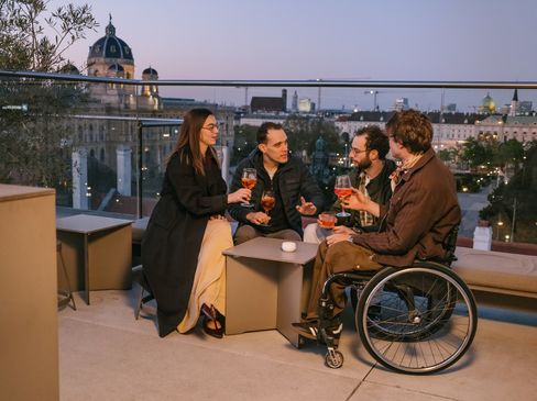 Vier Personen sitzen am Abend gemeinsam auf der MQ Libelle und unterhalten sich bei Getränken. Eine Person nutzt einen Rollstuhl, im Hintergrund ist die Wiener Skyline mit der Kuppel des Naturhistorischen Museums zu sehen.