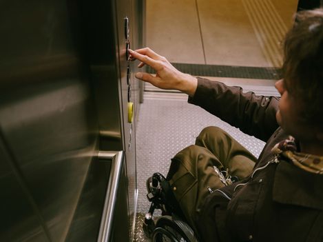 Eine Person im Rollstuhl betätigt die Bedienelemente eines barrierefreien Aufzugs. Die Aufnahme zeigt den Innenraum des Aufzugs mit gut erreichbaren Tasten.