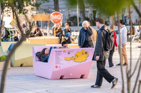 Besucher:innen sitzen und gehen im Hof des MuseumsQuartier; im Vordergrund ein rosa Enzi mit „Enzi on Tour“-Karte.