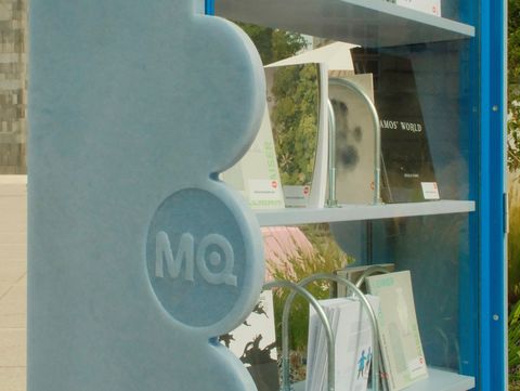 Blauer Bücherschrank mit Glasfront und mehreren Büchern auf drei Regalböden, links seitlich mit erhabenen Kreisen und den Buchstaben "MQ".