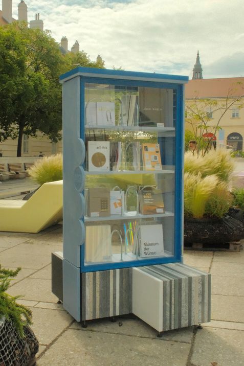 Blauer Bücherschrank mit Glasfront und Büchern auf mehreren Regalböden im Freien auf gepflastertem Platz mit Pflanzen und Gebäuden im Hintergrund.