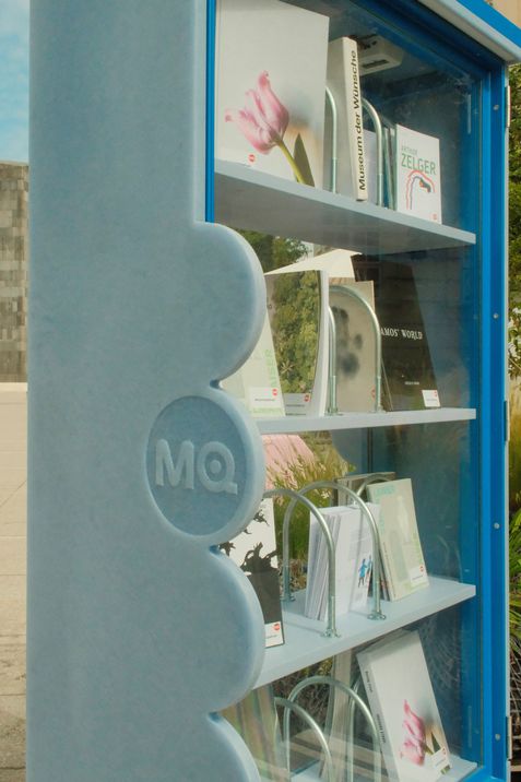 Blauer Bücherschrank mit Glasfront und mehreren Büchern auf drei Regalböden, links seitlich mit erhabenen Kreisen und den Buchstaben "MQ".