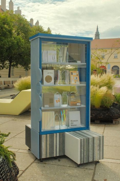 Blauer Bücherschrank mit Glasfront und Büchern auf mehreren Regalböden im Freien auf gepflastertem Platz mit Pflanzen und Gebäuden im Hintergrund.