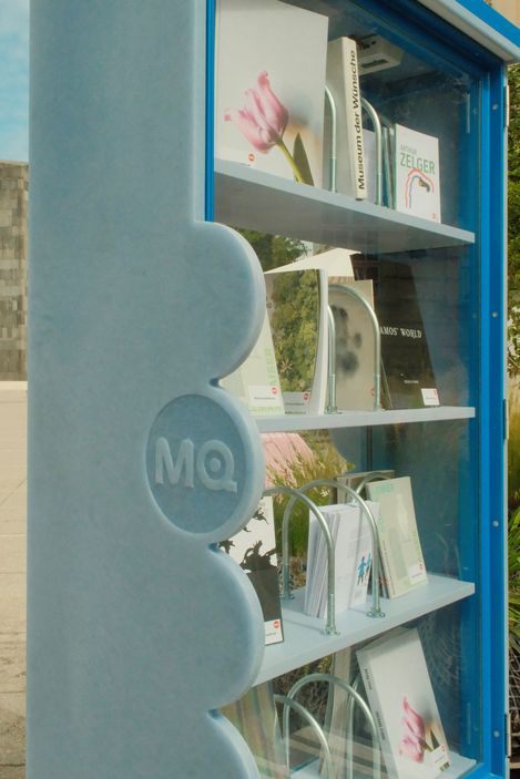 Blauer Bücherschrank mit Glasfront und mehreren Büchern auf drei Regalböden, links seitlich mit erhabenen Kreisen und den Buchstaben "MQ".