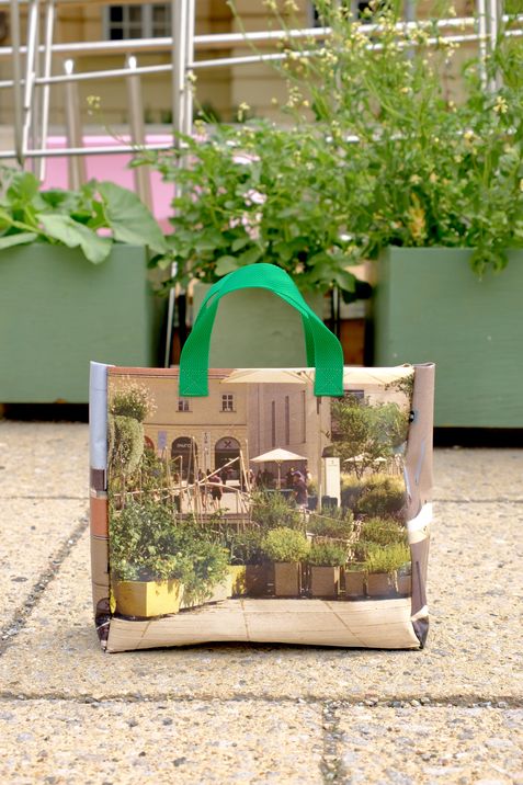 Kleine Tasche mit grünem Henkel und Fotodruck eines Innenhofs mit Pflanzen und Sitzgelegenheiten auf Steinboden vor grünen Pflanzkästen.