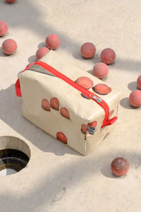Rechteckige Tasche mit rotem Reißverschluss und Aufdruck von Pfirsichen auf hellem Untergrund, umgeben von mehreren kleinen Pfirsichkugeln auf einer hellen Fläche mit einem runden Loch.