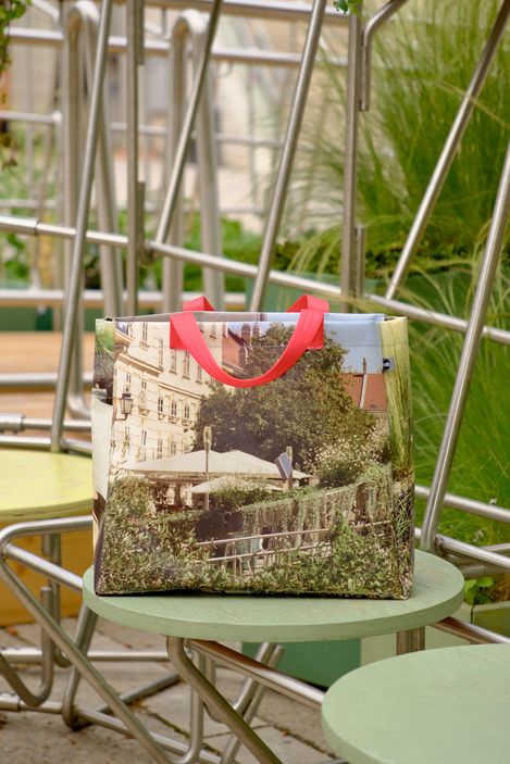 Tasche mit Fotodruck einer urbanen Szene mit Gebäuden, Bäumen und einem Straßenschild, roter Henkel, auf grünem runden Tisch vor Metallstühlen.