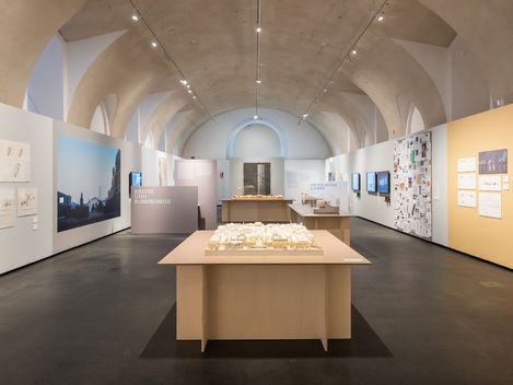 Ausstellungsraum mit hohen gewölbten Decken, mehreren Ausstellungstafeln an den Wänden und einem Modell auf einem Tisch in der Raummitte