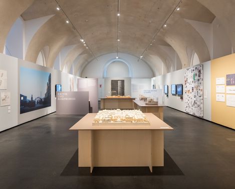 Ausstellungsraum mit hohen gewölbten Decken, mehreren Ausstellungstafeln an den Wänden und einem Modell auf einem Tisch in der Raummitte