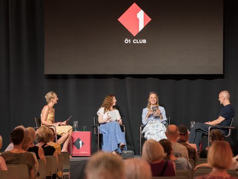 Vier Personen sitzen auf einer Bühne vor Publikum, drei Frauen und ein Mann, mit einem Bildschirm im Hintergrund, der ein rotes Logo mit der Aufschrift 'Ö1 CLUB' zeigt