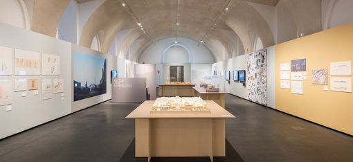 Ausstellungsraum mit hohen gewölbten Decken, mehreren Ausstellungstafeln an den Wänden und einem Modell auf einem Tisch in der Raummitte