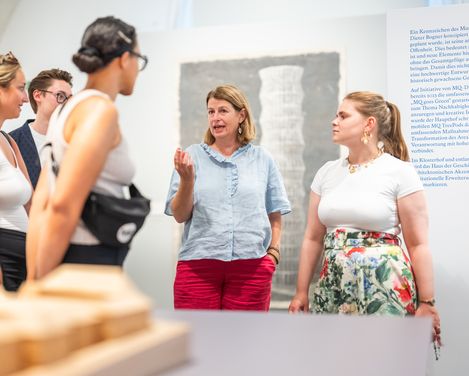 Fünf Personen in einem Ausstellungsraum, eine Frau spricht und gestikuliert, andere hören zu, im Vordergrund unscharfe Ausstellungsexponate
