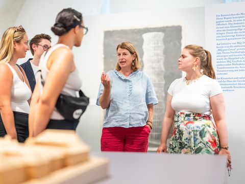 Fünf Personen in einem Ausstellungsraum, eine Frau spricht und gestikuliert, andere hören zu, im Vordergrund unscharfe Ausstellungsexponate