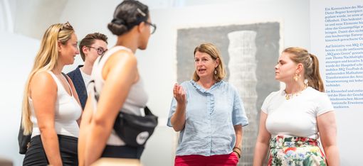 Fünf Personen in einem Ausstellungsraum, eine Frau spricht und gestikuliert, andere hören zu, im Vordergrund unscharfe Ausstellungsexponate