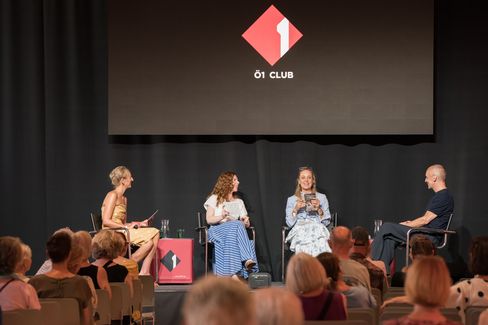 Vier Personen sitzen auf einer Bühne vor Publikum, drei Frauen und ein Mann, mit einem Bildschirm im Hintergrund, der ein rotes Logo mit der Aufschrift 'Ö1 CLUB' zeigt