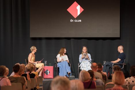 Vier Personen sitzen auf einer Bühne vor Publikum, drei Frauen und ein Mann, mit einem Bildschirm im Hintergrund, der ein rotes Logo mit der Aufschrift 'Ö1 CLUB' zeigt