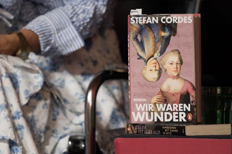 Buch mit dem Titel 'Wir waren Wunder' von Stefan Cordes steht auf einem Stapel Bücher neben einer Person mit gestreiftem Hemd und Rollstuhl