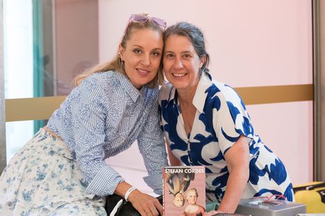 Zwei Frauen sitzen nah beieinander, eine trägt eine gestreifte Bluse und einen Rock mit floralem Muster, die andere ein weißes Kleid mit blauem Muster, vor ihnen steht ein Buch mit dem Titel 'Stefan Cordes'