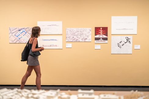 Frau in kurzem Kleid und Rucksack betrachtet Architekturzeichnungen an einer hellen Wand in einer Ausstellung