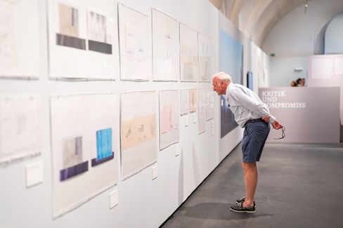 Älterer Mann beugt sich vor und betrachtet Kunstwerke an einer weißen Ausstellungswand in einem hellen Raum mit gewölbter Decke