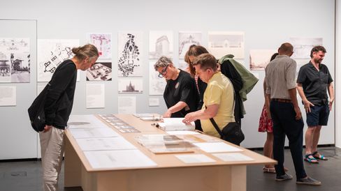 Mehrere Personen betrachten auf einem Tisch in einer Galerie oder Ausstellung liegende Zeichnungen und Pläne, im Hintergrund hängen weitere Bilder an der Wand.