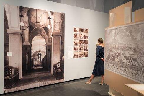 Eine Frau in blauem Kleid betrachtet historische Fotografien und Zeichnungen in einer Ausstellung mit hohen Wänden und gedämpfter Beleuchtung