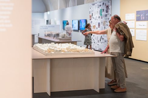 Zwei ältere Personen betrachten ein Architekturmodell auf einem Tisch in einer Ausstellung mit weiteren Besuchern und ausgestellten Bildern im Hintergrund