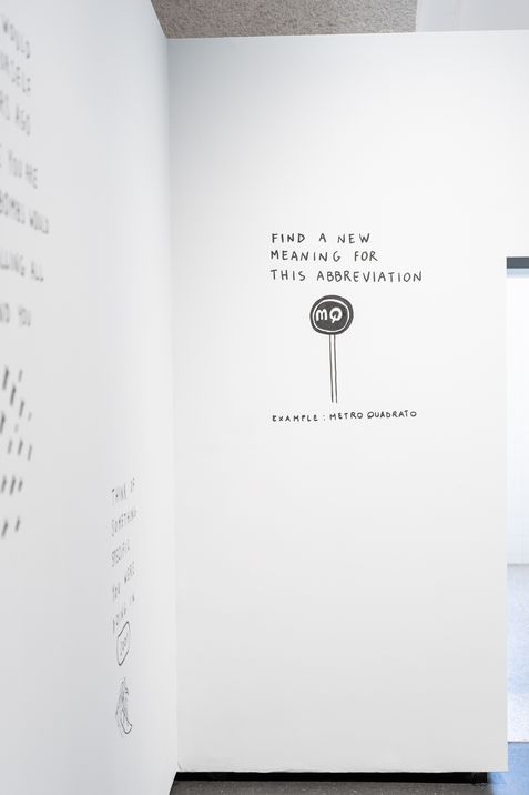 Weiße Wand mit schwarzer Schrift und einer Zeichnung eines runden Schilds mit den Buchstaben 'mq' und dem Text 'FIND A NEW MEANING FOR THIS ABBREVIATION EXAMPLE: METRO QUADRATO'